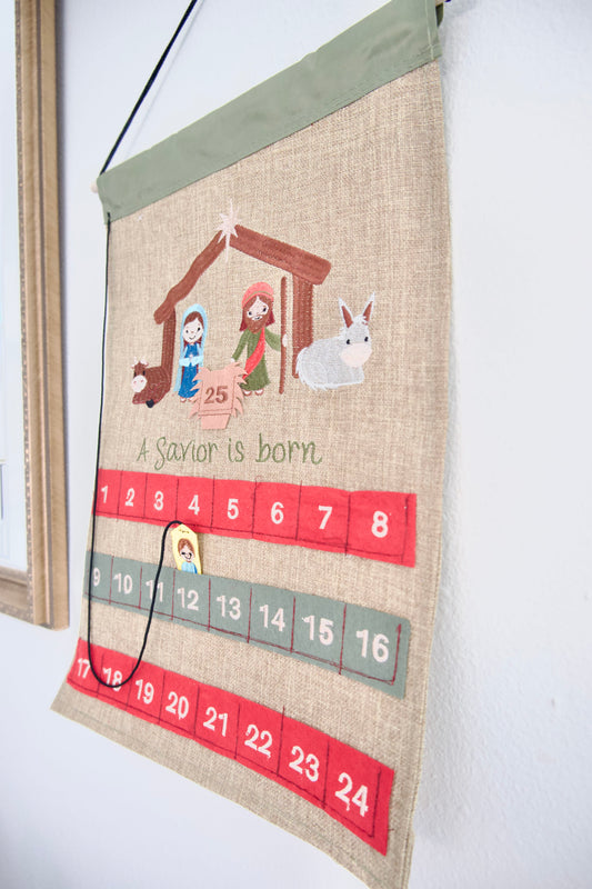 A Savior Is Born fabric Advent calendar with embroidered Nativity scene, numbered pockets, Baby Jesus piece, and 25-day Christmas countdown